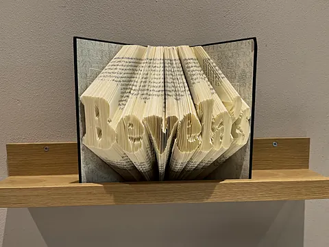 Gefaltetes Buch mit dem Schriftzug Bevela's auf einem Holzregal